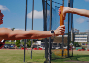 Two athletes exchange an orange relay baton during a handoff on a sunny track field.