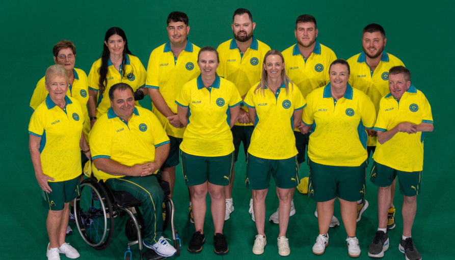 Group portrait of a mixed-gender sports team in yellow and green uniforms posing on a green floor; one person in a wheelchair is in the front row left.