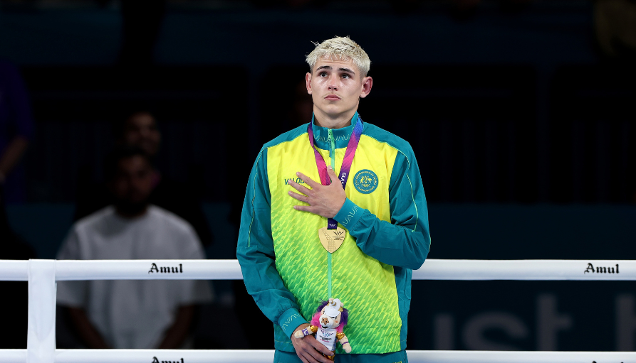 Young athlete in a bright teal and lime jacket with a gold medal, hand over heart, standing in a boxing ring and holding a plush toy.
