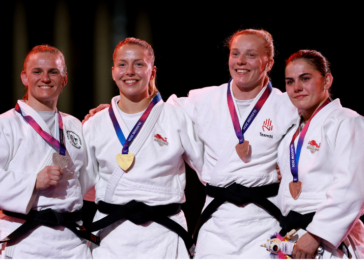 Four female judo athletes on a podium, wearing white judogi with black belts, each with medals around their necks and smiling with arms around each other.