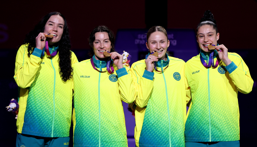 Four female athletes in bright neon jackets bite medals while posing on stage, celebrating a podium finish with smiles and blue-green outfits.