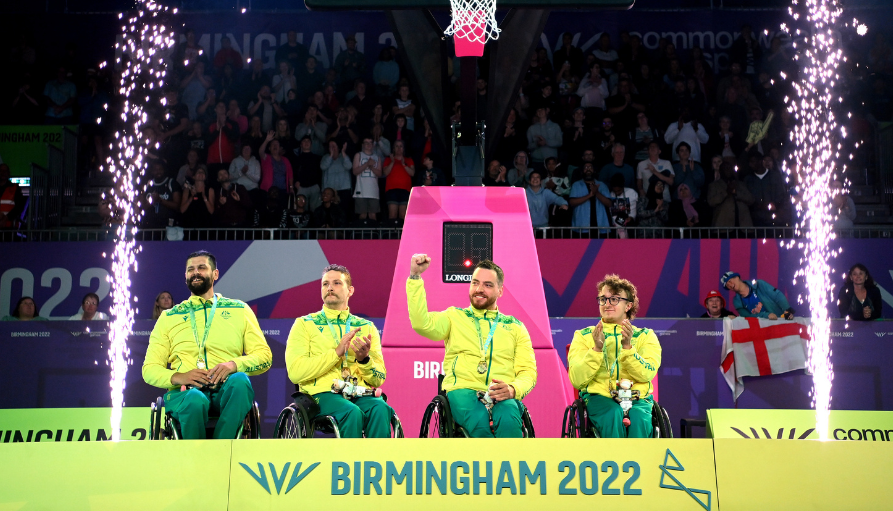 Paralympic medalists in wheelchairs on a podium celebrate with medals as fireworks erupt, Birmingham 2022 backdrop behind them.