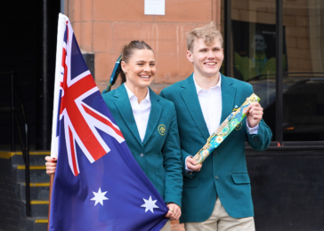 Two students in teal blazers smile while holding an Australian flag outside a brick building, with a decorative gift wrap roll in hand.