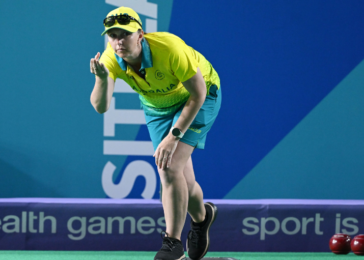 Female athlete in a yellow Australian shirt and blue shorts crouches to release a bowls ball on a green court, with 'Sport is' banners in the background.