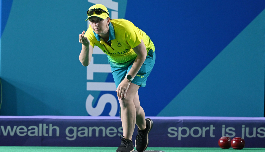 Female athlete in a yellow Australian shirt and blue shorts crouches to release a bowls ball on a green court, with 'Sport is' banners in the background.