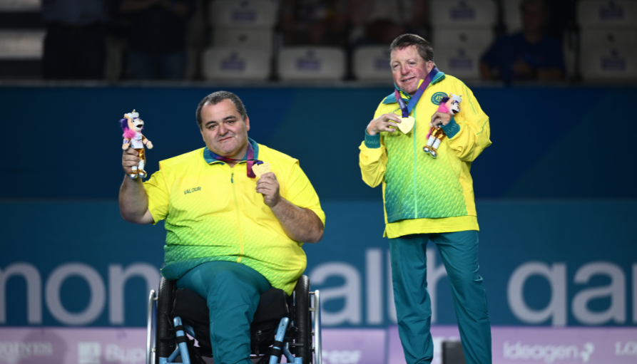 Two para-athletes in neon jackets pose on stage; one in a wheelchair holds a mascot while the other shows a medal and trophy against a blue backdrop.