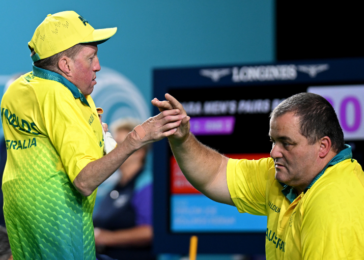 Two athletes in yellow/green uniforms shake hands on a sports arena, with a scoreboard behind them.