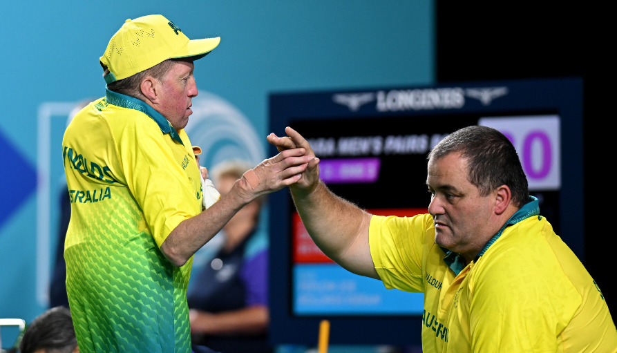 Two athletes in yellow/green uniforms shake hands on a sports arena, with a scoreboard behind them.
