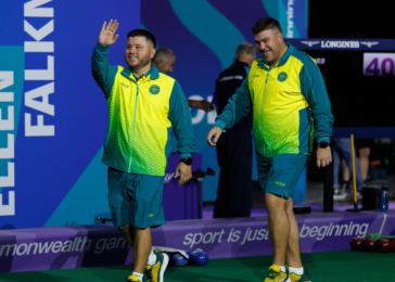 Two male athletes in teal and lime-green jackets walk onto a lawn bowls area, waving to spectators with a scoreboard behind them at a Commonwealth Games venue.
