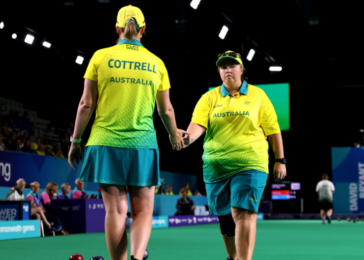 Two Australian athletes in yellow-green uniforms shake hands on a green indoor sports court after a match, with bowls visible on the floor and spectators in the background.