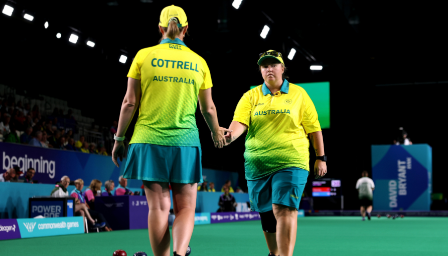 Two Australian athletes in yellow-green uniforms shake hands on a green indoor sports court after a match, with bowls visible on the floor and spectators in the background.