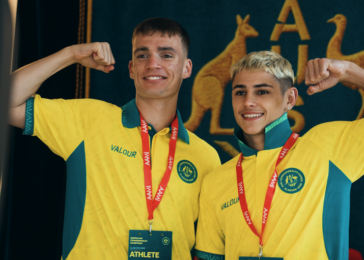 Two athletes in bright yellow jerseys flex their biceps and smile for a photo, each wearing a red lanyard with a medal against a dark blue backdrop.