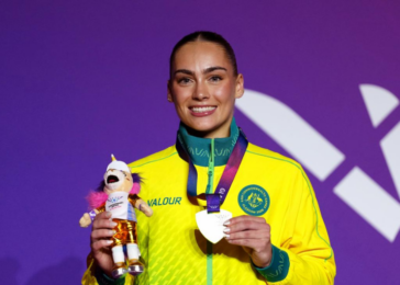 Female athlete in a bright yellow jacket smiles on a purple backdrop, holding a plush mascot trophy and a medal around her neck.
