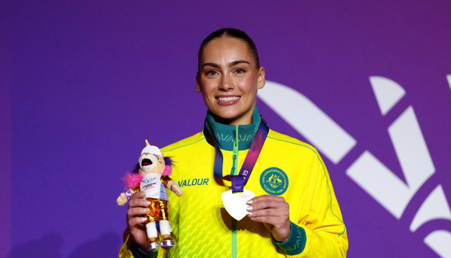 Female athlete in a bright yellow jacket smiles on a purple backdrop, holding a plush mascot trophy and a medal around her neck.