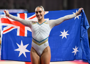 Gymnast in a white leotard with gold accents holds an Australian flag wide, smiling on stage.