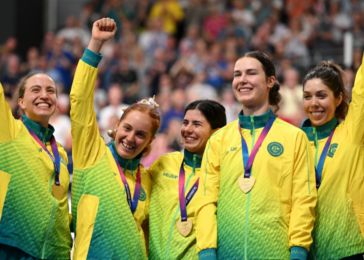 Five female athletes in bright yellow-green jackets celebrate on a podium with medals and raised fists.