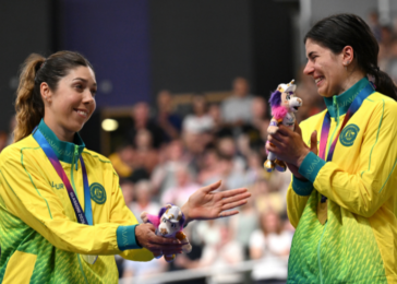 Two female athletes in yellow-green uniforms high-five on a podium, each holding a small mascot plush toy to celebrate.