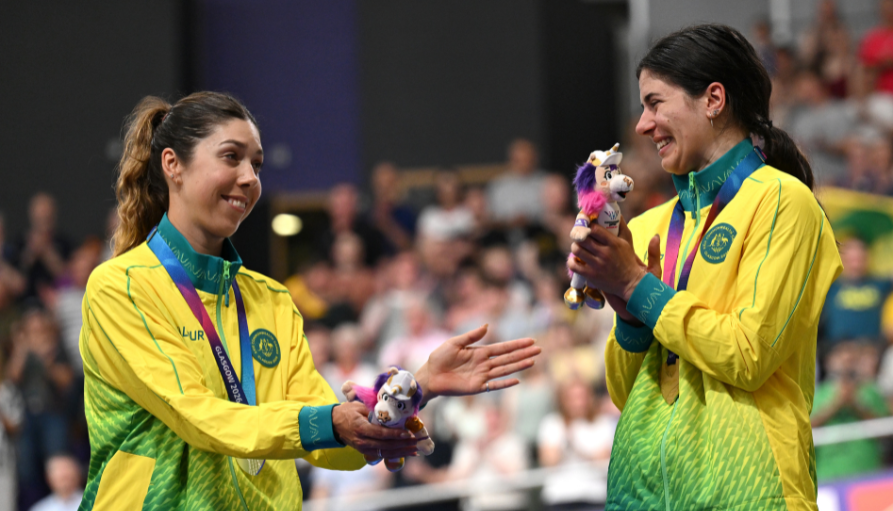 Two female athletes in yellow-green uniforms high-five on a podium, each holding a small mascot plush toy to celebrate.