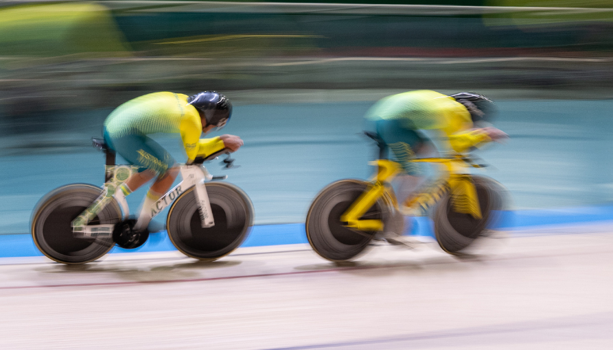 Two cyclists in teal and yellow race on a velodrome, motion blurred as they sprint by a water backdrop.