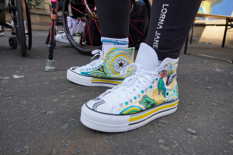 White high-top Converse sneakers with a bright teal, yellow, and green Paisley-style pattern on the toes, worn with black leggings, foreground on pavement.