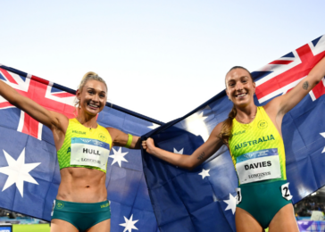 Two Australian track athletes celebrate victory, holding the national flag between them and smiling on a stadium track surface.