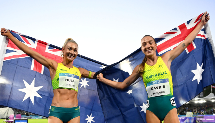 Two Australian track athletes celebrate victory, holding the national flag between them and smiling on a stadium track surface.