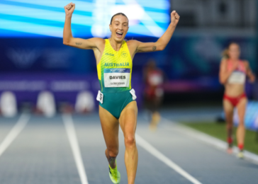 Australian sprinter crosses finish line with arms raised in victory, smiling on a blue track with a neon-yellow and green uniform.