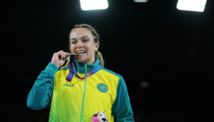 Female athlete in teal and neon yellow jacket biting a gold medal, smiling at the camera.
