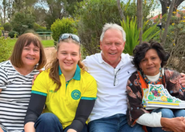 Four people sit on a park bench, smiling at the camera; a woman in a yellow polo sits second from the left, flanked by an older man in a white shirt and two other women. The rightmost woman holds a colorful book or binder.