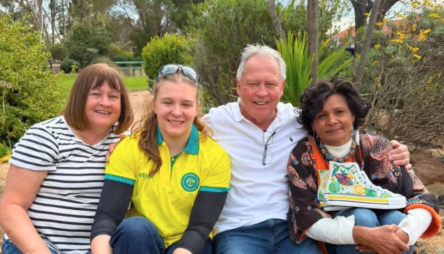Four people sit on a park bench, smiling at the camera; a woman in a yellow polo sits second from the left, flanked by an older man in a white shirt and two other women. The rightmost woman holds a colorful book or binder.