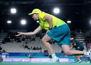 Athlete in neon yellow shirt and blue shorts lunges to release a ball on an indoor sports court with spectators in the background.