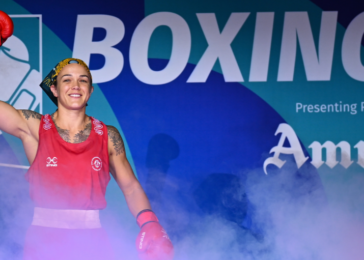 Female boxer in red gear raises a gloved fist in victory on a boxing event stage with a blue backdrop that reads 'BOXING'.