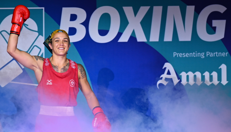 Female boxer in red gear raises a gloved fist in victory on a boxing event stage with a blue backdrop that reads 'BOXING'.