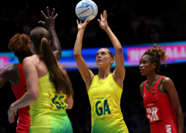 Female netball players in yellow and red uniforms contest the ball mid-air on a brightly lit court, GA reaching for a pass or shot.