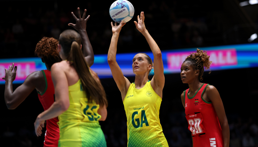 Female netball players in yellow and red uniforms contest the ball mid-air on a brightly lit court, GA reaching for a pass or shot.