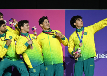 A team of five male athletes in yellow-green uniforms posing on stage with medals, taking a selfie with a phone held by the rightmost teammate.