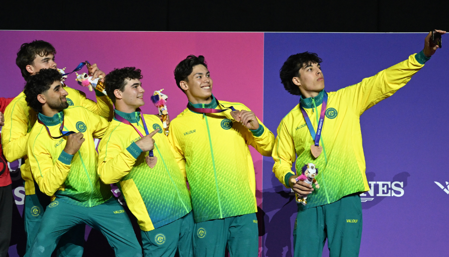 A team of five male athletes in yellow-green uniforms posing on stage with medals, taking a selfie with a phone held by the rightmost teammate.