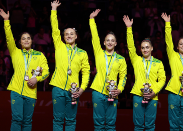 Five female athletes in bright yellow jackets and teal pants, each with medals, wave on stage while holding trophies.