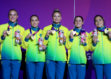 Five female athletes in neon jackets hold medals and mascot plush toys on stage with a purple background, smiling proudly.