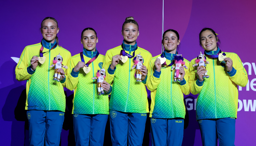 Five female athletes in neon jackets hold medals and mascot plush toys on stage with a purple background, smiling proudly.