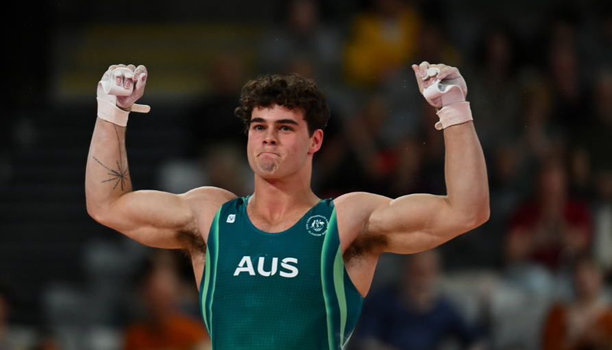 Male athlete in a teal Australia singlet raises both arms in victory, fists clenched, wearing white wrist wraps and a visible star tattoo on his left arm.