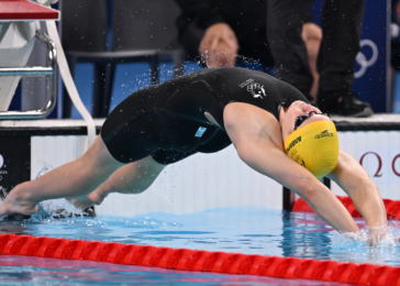 Swimmer wearing a black suit and yellow cap diving into a pool at the start of a race, creating a splash near the starting block.
