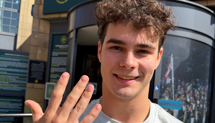 Young man with curly brown hair smiles at the camera while waving his hand outdoors near a building entrance.