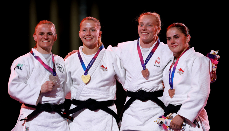 Four female judo athletes in white judogis with black belts, posing on stage with medals around their necks.