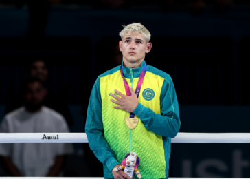 Young male athlete in teal and lime-green uniform stands in a ring, hand on chest, wearing medals and a plush trophy.