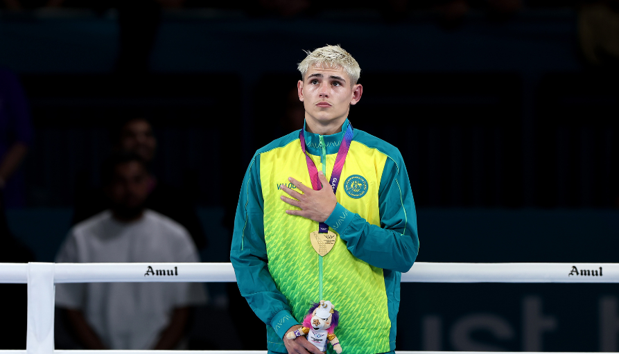 Young male athlete in teal and lime-green uniform stands in a ring, hand on chest, wearing medals and a plush trophy.