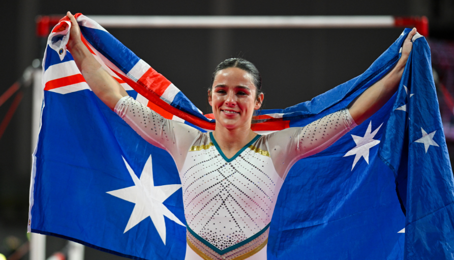 Gymnast stands with arms raised, holding an Australian flag behind her, smiling after a routine fall or victory moment.
