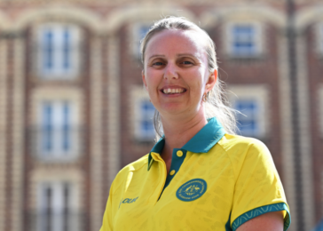 Smiling woman in a yellow polo shirt with teal collar, standing outdoors in front of a brick campus building.