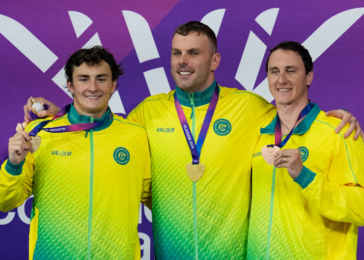 Three male athletes in yellow–green jackets on a podium, each holding a medal and smiling against a purple backdrop.