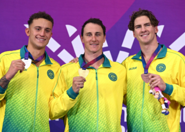Three male athletes in matching yellow-green jackets pose with their medals against a purple backdrop (left silver, center gold, right bronze).